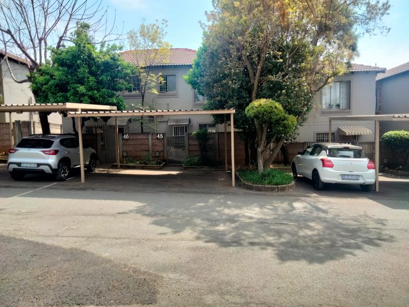 Front exterior view of a townhouse complex featuring covered parking bays, brick boundary walls, and established trees.