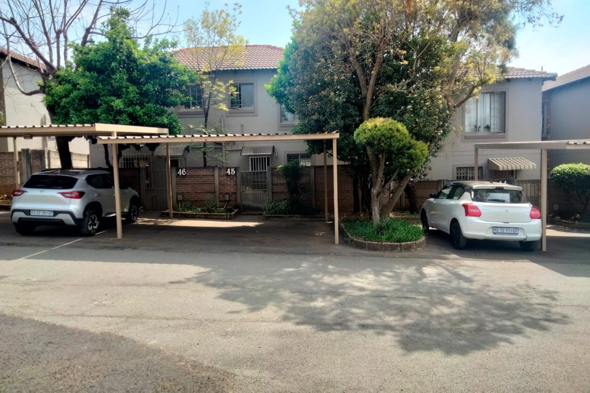 Front exterior view of a townhouse complex featuring covered parking bays, brick boundary walls, and established trees.