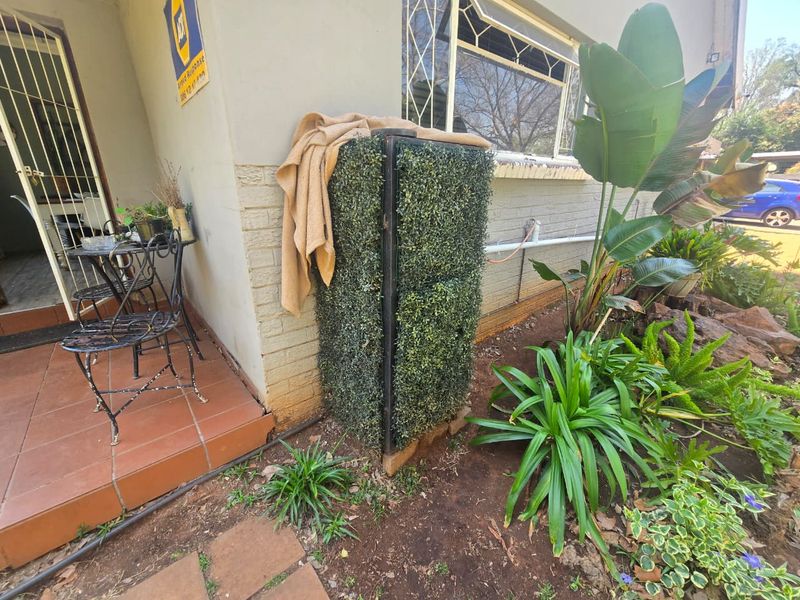 A covered patio area featuring tiled flooring, a security gate, and a window with burglar bars, adjacent to a small garden bed.