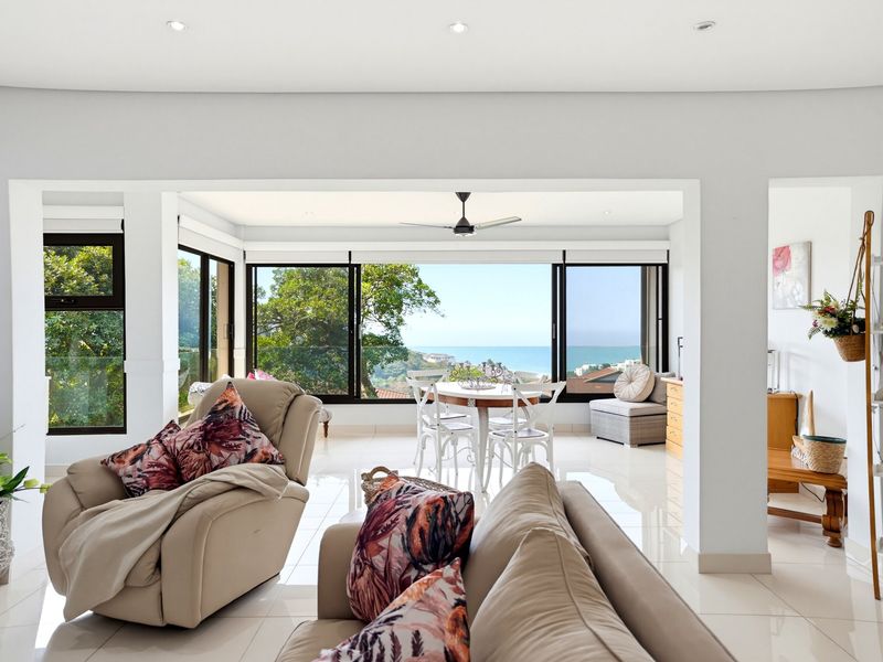 Bright open-plan living and dining area featuring large sliding doors that open to a scenic ocean view, complemented by modern tiled floors and recessed lighting.