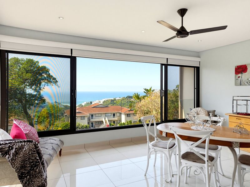 Bright dining area featuring large sliding glass doors that offer expansive ocean and coastal views, complemented by modern tiled flooring and a ceiling fan.