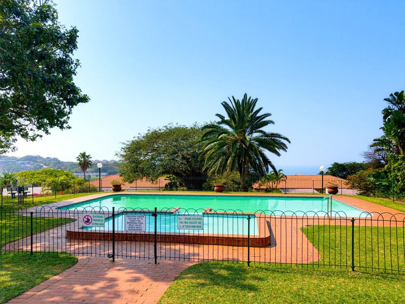 A well-maintained communal swimming pool area featuring a main pool, a shallow wading pool, brick paving, and secure metal perimeter fencing.