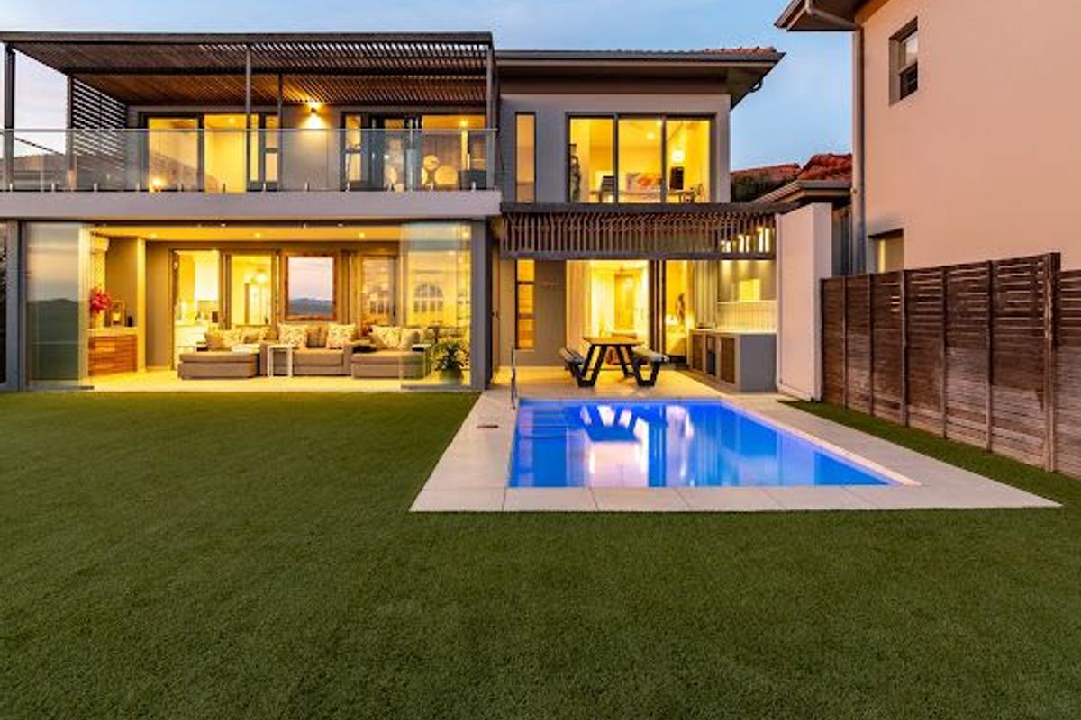 Rear view of a modern double-story home featuring a swimming pool, artificial lawn, covered patio, and an upper-level balcony with timber pergola.