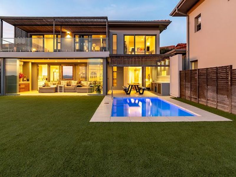 Rear view of a modern double-story home featuring a swimming pool, artificial lawn, covered patio, and an upper-level balcony with timber pergola.