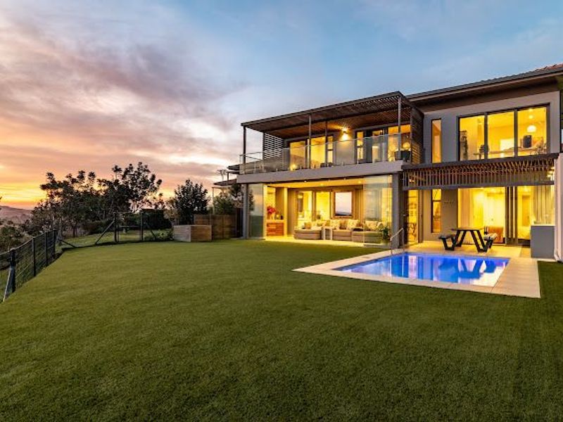 Modern double-story residence featuring a rear patio, balcony with glass balustrades, and a swimming pool overlooking a landscaped garden at sunset.