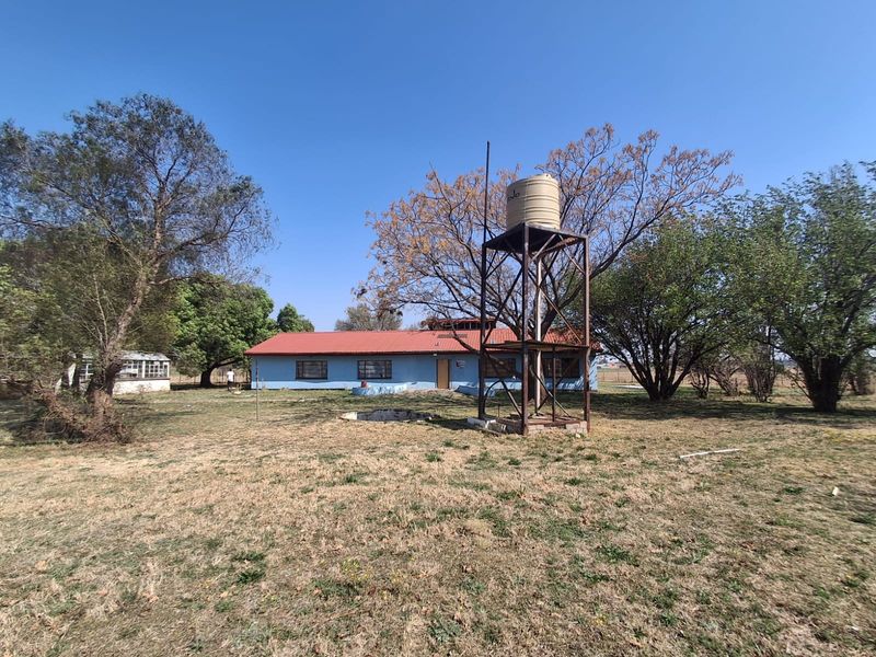 Rear view of a farm dwelling featuring a metal water tank stand, corrugated roof, and expansive surrounding grounds.