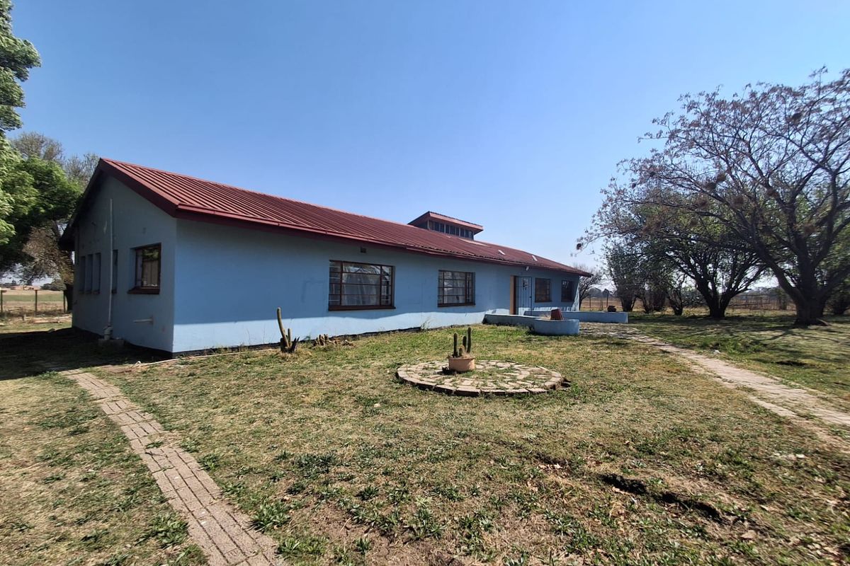 Rear view of a single-story farmhouse featuring a corrugated metal roof, masonry walls, and surrounding garden space with brick pathways.