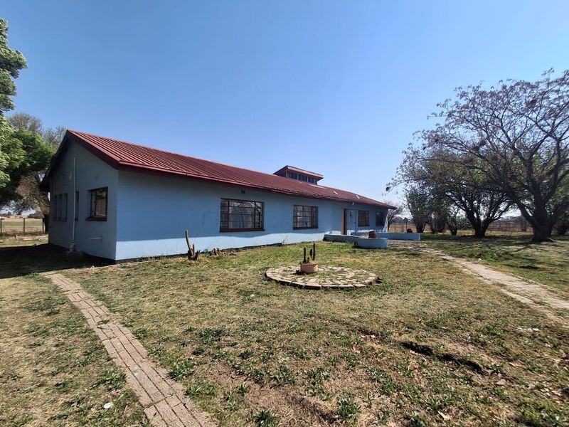 Rear view of a single-story farmhouse featuring a corrugated metal roof, masonry walls, and surrounding garden space with brick pathways.