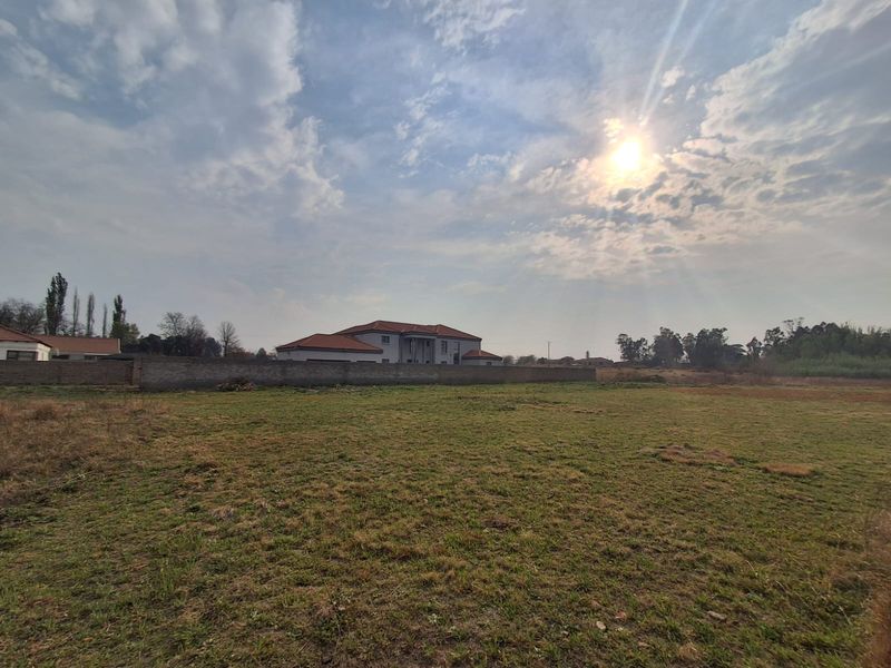 A wide-angle view of a vacant residential plot featuring natural grassland and a perimeter boundary wall with neighboring properties in the background.