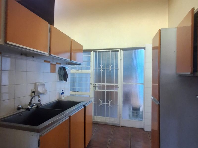 A dated kitchen featuring exposed timber ceiling beams, tiled flooring, double basin sink, and original cabinetry with direct access to an exterior security door.