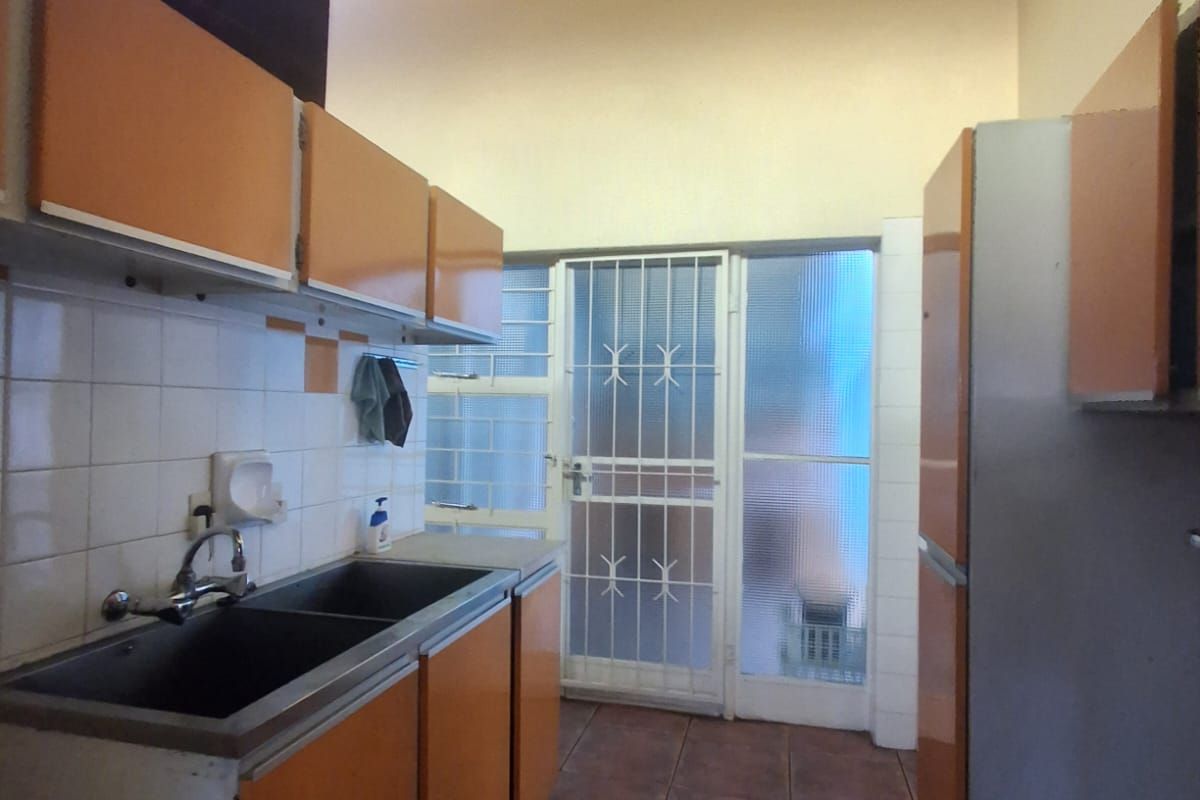 A dated kitchen featuring exposed timber ceiling beams, tiled flooring, double basin sink, and original cabinetry with direct access to an exterior security door.