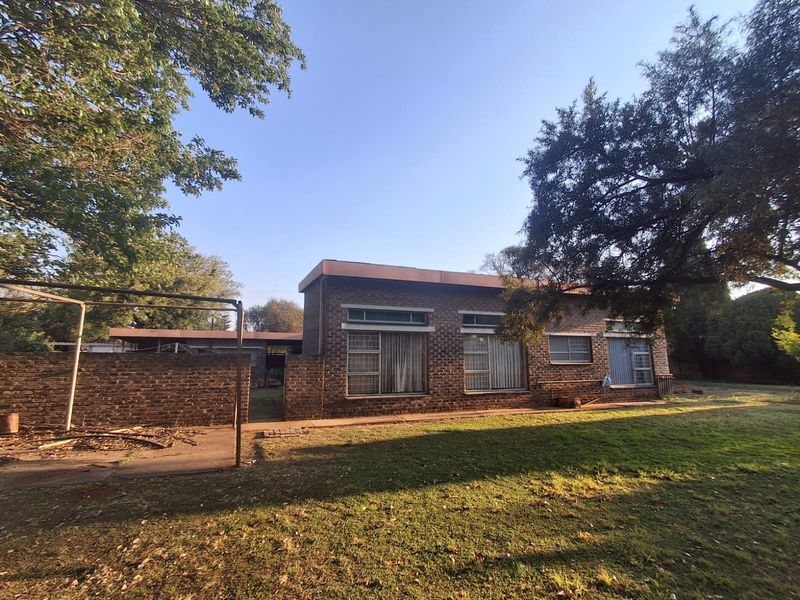 Rear view of a single-story face brick house featuring metal window frames, a concrete patio area, and a large garden with mature trees.