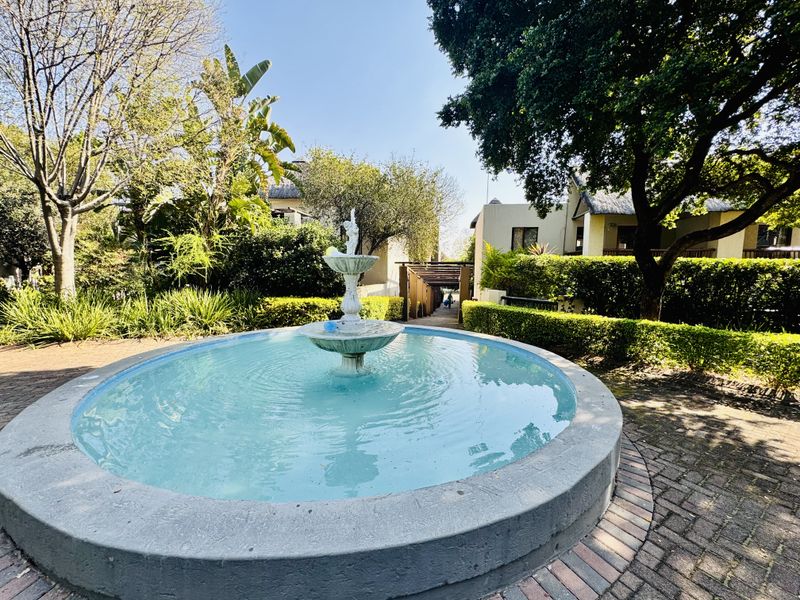 A well-maintained garden area featuring a central circular water fountain, brick paved pathways, and mature landscaping with a wooden pergola in the background.