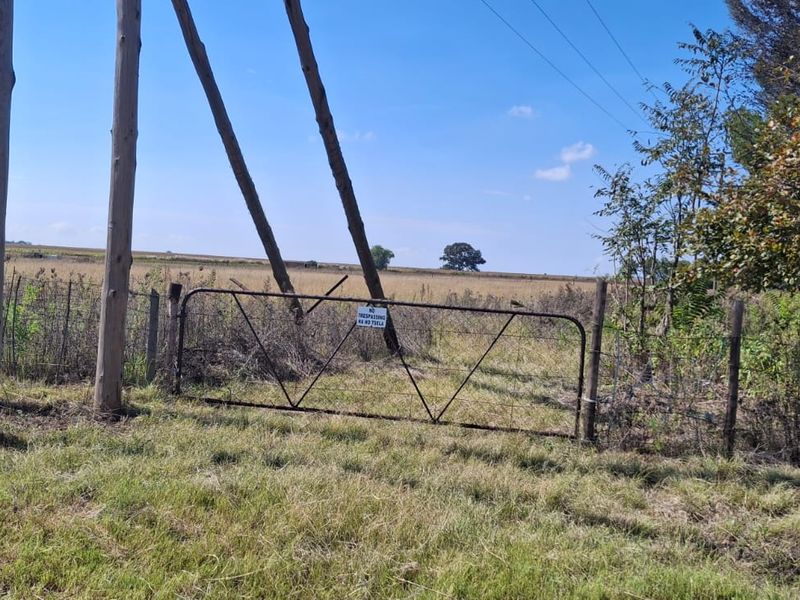 A view of a rural farm entrance featuring a metal gate, wire fencing, and wooden utility poles set against an open field.