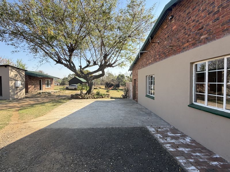 Rear exterior view of the small holding featuring exposed brick walls, concrete driveway, and established trees on the property grounds.