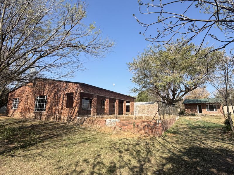Rear view of a face-brick small holding property featuring a brick retaining wall, mature trees, and a secondary outbuilding structure.