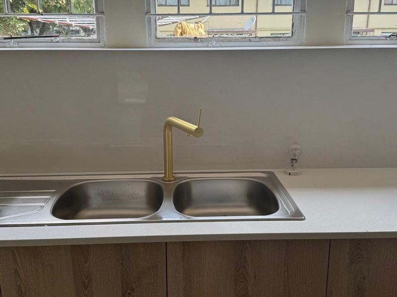 A clean kitchen area featuring a modern brass faucet, stainless steel double sink, and light wood-finish cabinetry under large windows with security bars.