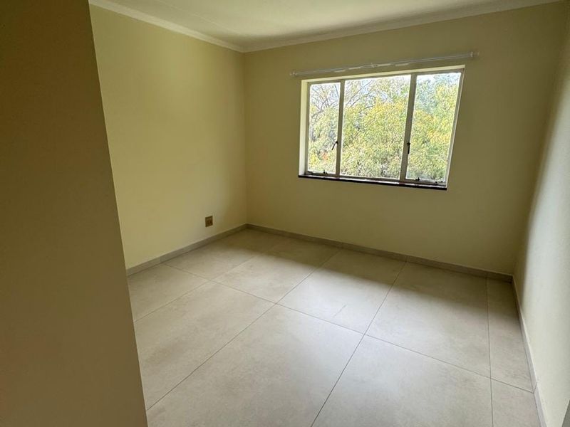 A bright, empty bedroom featuring neutral tiled flooring, clean walls, and a large window providing natural light and a pleasant outdoor view.