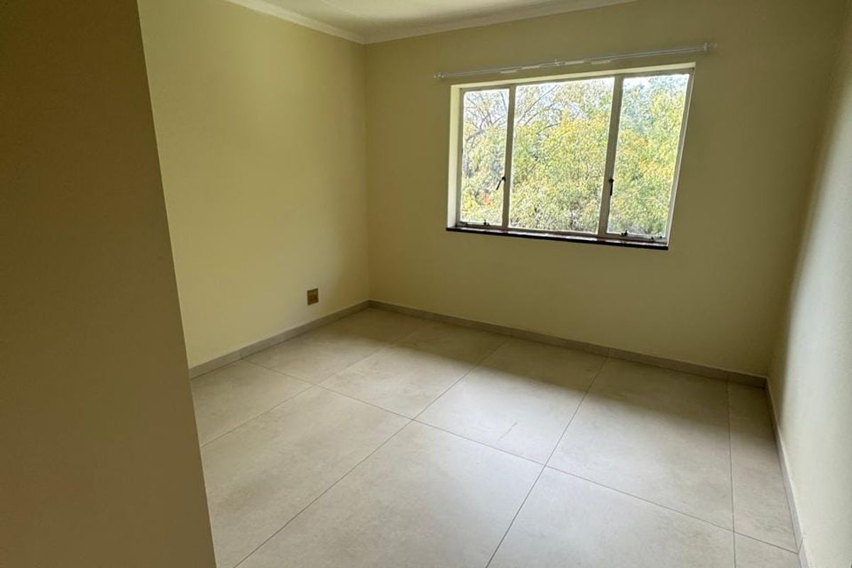 A bright, empty bedroom featuring neutral tiled flooring, clean walls, and a large window providing natural light and a pleasant outdoor view.