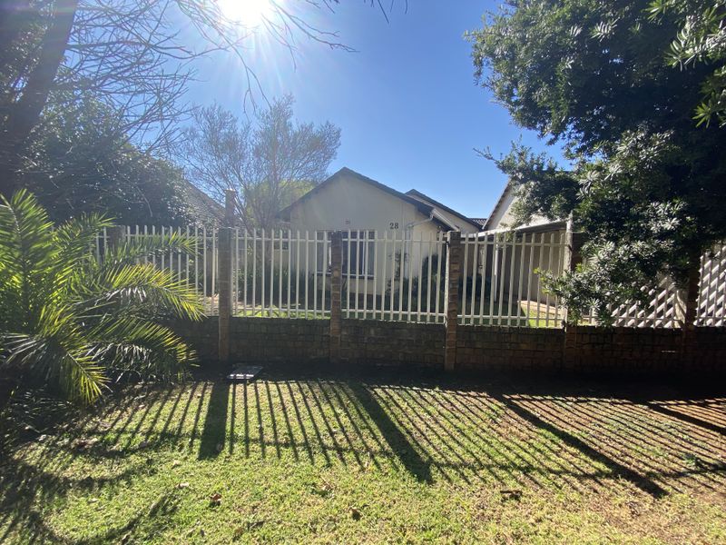 Front view of the property featuring a secure brick and metal picket fence, paved driveway, and established garden area.