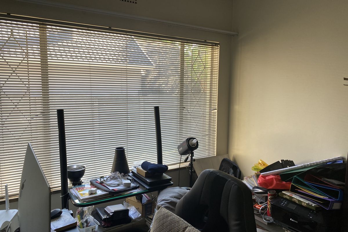 A bedroom space featuring a large window with venetian blinds and a ceiling air vent, currently utilized as a home office area.