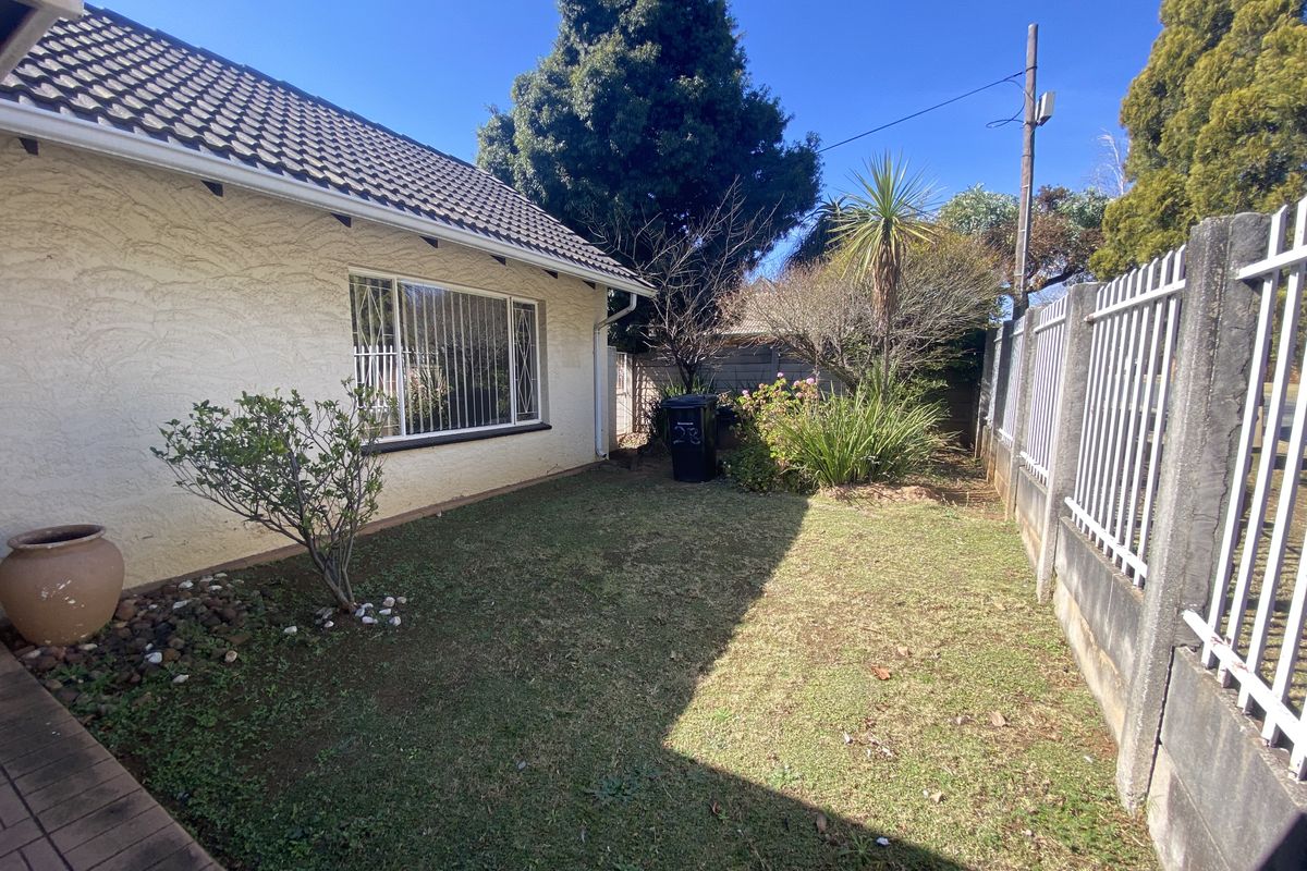 A rear garden area featuring a lawn, boundary wall with metal fencing, and security bars on the house windows.