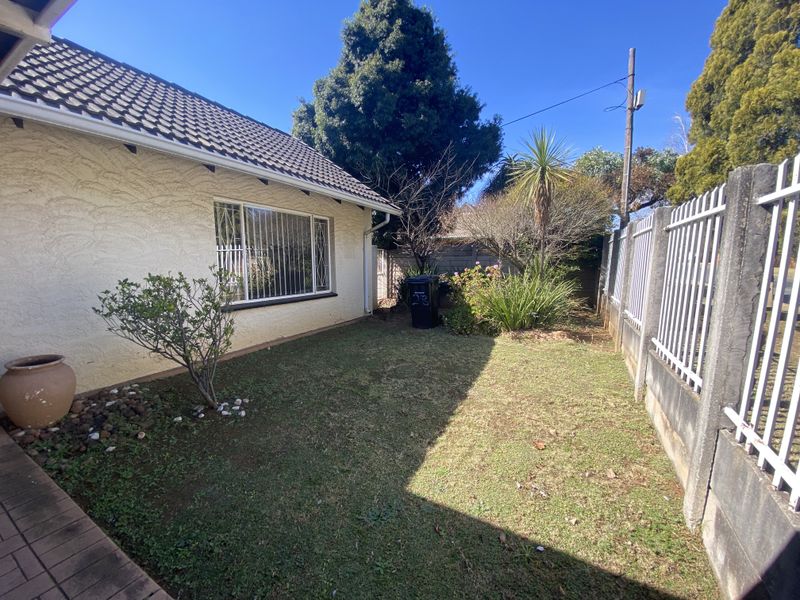 A rear garden area featuring a lawn, boundary wall with metal fencing, and security bars on the house windows.
