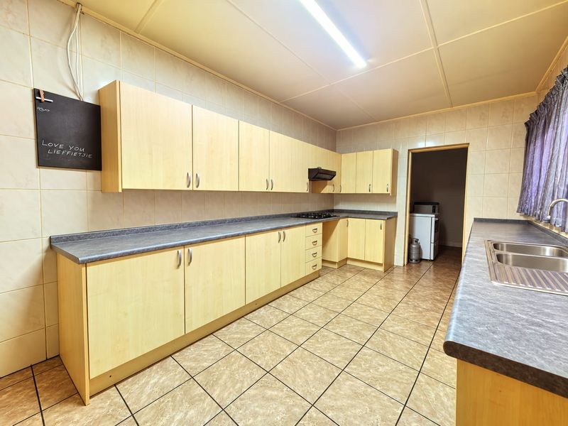 Functional kitchen featuring light wood-finish cabinetry, laminate countertops, a double stainless steel sink, built-in hob, and tiled flooring throughout.