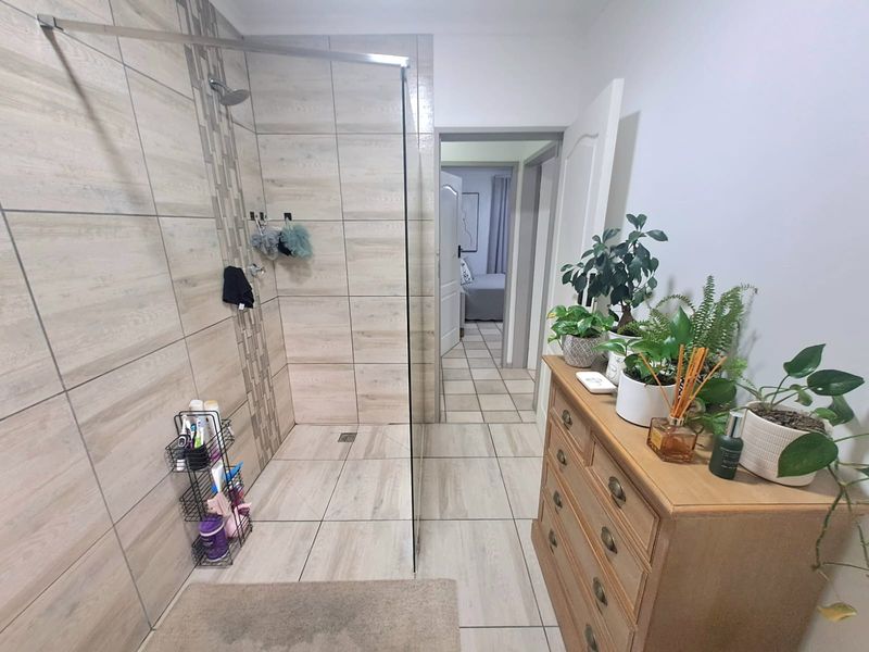 A modern bathroom featuring a spacious walk-in shower with neutral wood-look tiling and a glass screen, leading into an adjacent bedroom.