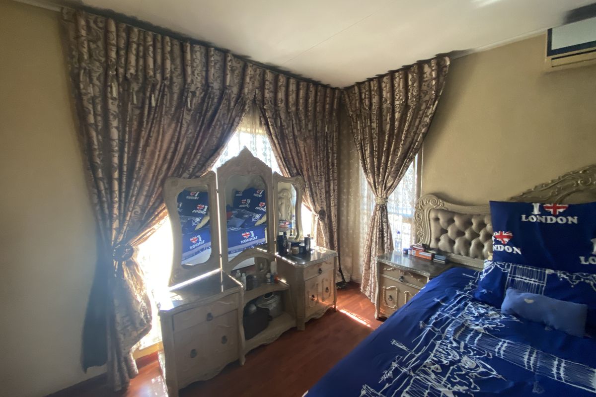 A bedroom featuring laminate flooring and a wall-mounted air conditioning unit, with large windows dressed in heavy decorative curtains.