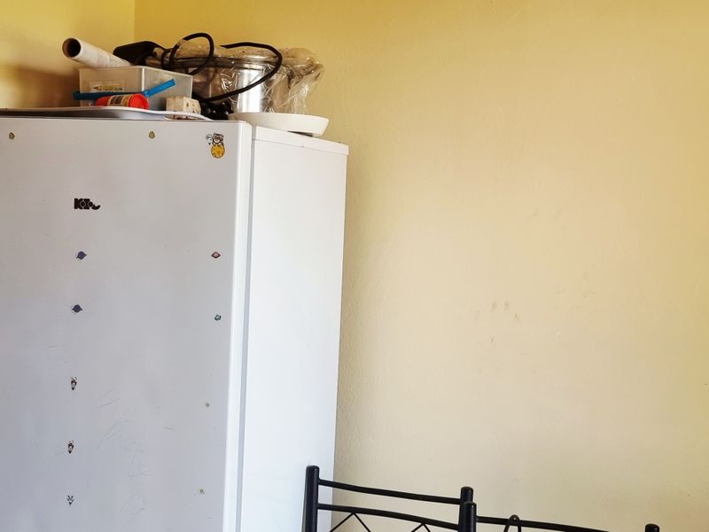 A close-up view of a corner showing a refrigerator and a plain yellow wall with visible surface marks.