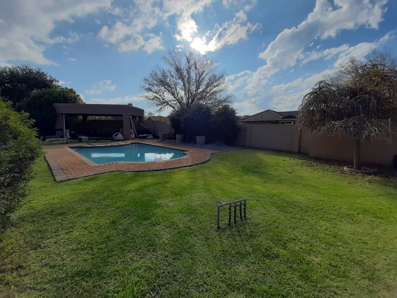A spacious backyard featuring a swimming pool with brick coping, a covered patio area, and a large lawn enclosed by boundary walls.
