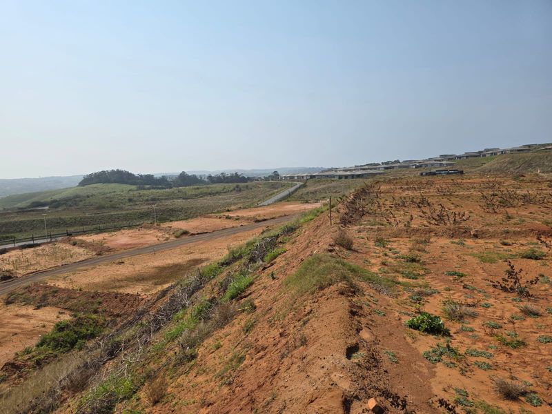 A wide-angle view of a vacant residential plot featuring natural terrain, earthworks, and surrounding landscape views in a developing estate.