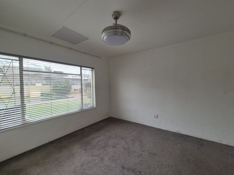 An empty bedroom featuring a ceiling fan, carpeted flooring, and large windows with venetian blinds, offering a blank canvas for renovation.