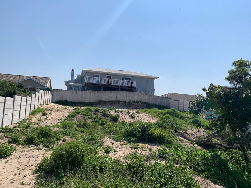 A view of a vacant residential plot featuring sandy terrain, native vegetation, and a precast concrete boundary wall with a neighboring house in the background.