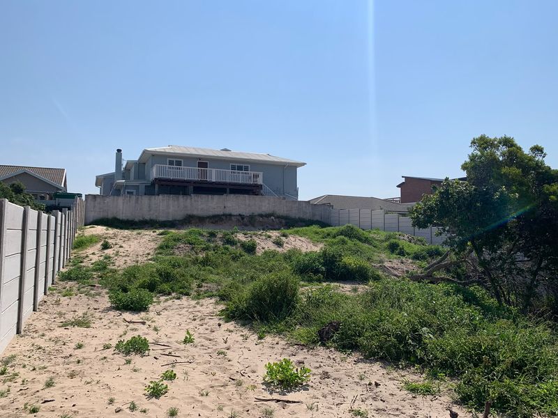 A view of a vacant residential plot featuring sandy terrain, coastal vegetation, and a precast concrete boundary wall with an adjacent property visible.