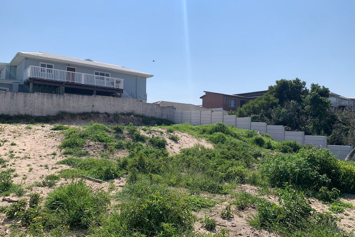 A view of a vacant residential plot featuring a sandy landscape, natural vegetation, and a concrete boundary wall with an elevated neighboring property.