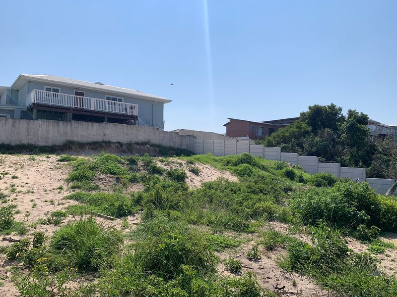 A view of a vacant residential plot featuring a sandy landscape, natural vegetation, and a concrete boundary wall with an elevated neighboring property.
