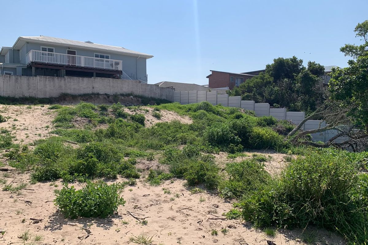A view of a vacant residential plot featuring sandy terrain, natural vegetation, and a precast concrete boundary wall with visible soil erosion.