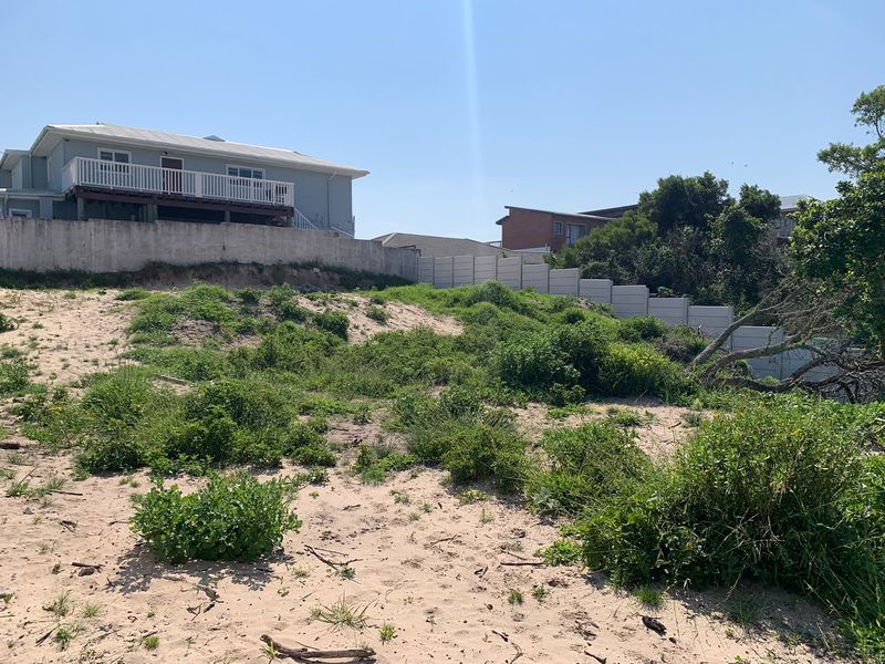 A view of a vacant residential plot featuring sandy terrain, natural vegetation, and a precast concrete boundary wall with visible soil erosion.