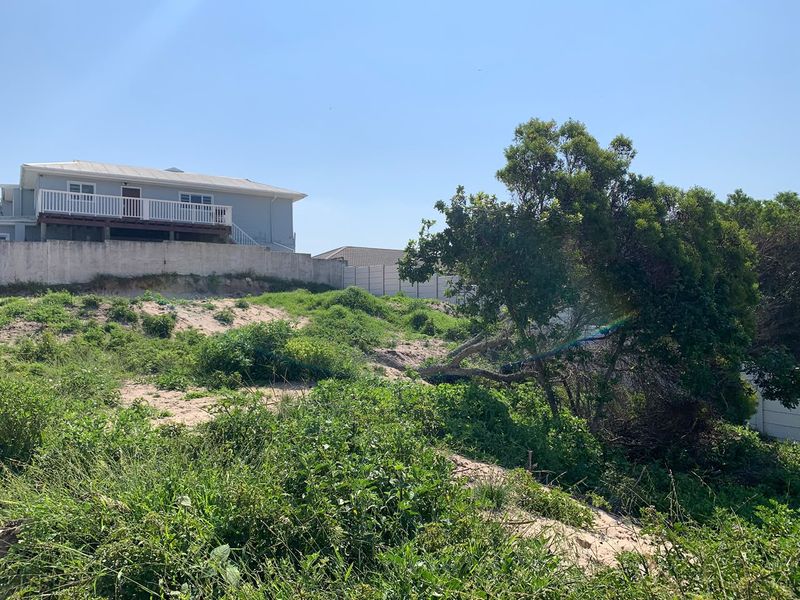 A view of a vacant residential plot featuring natural vegetation, a precast concrete boundary wall, and an adjacent elevated property with a timber deck.