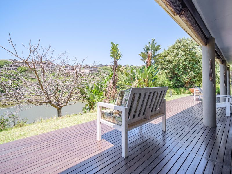 A spacious covered timber deck patio overlooking a tranquil pond and lush garden, featuring structural support columns and integrated outdoor blinds.