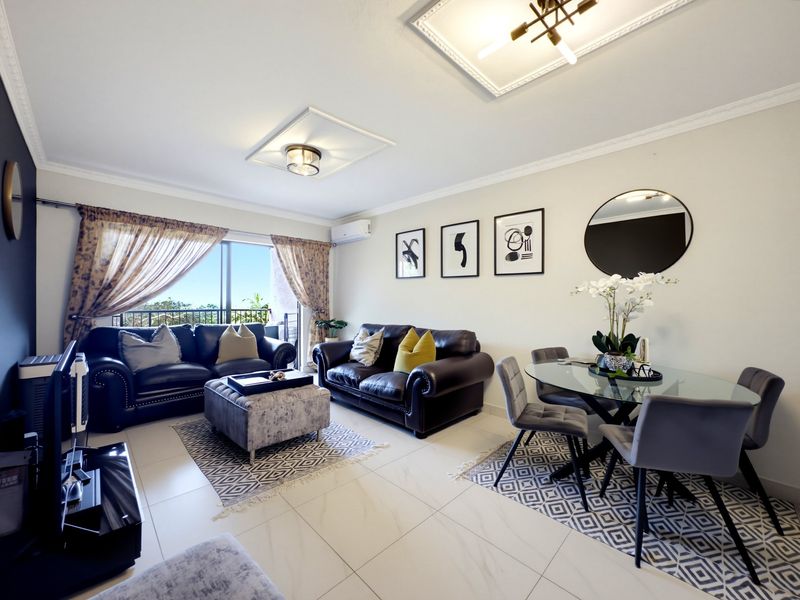 A bright open-plan living and dining area featuring tiled floors, decorative ceiling panels, and direct access to a balcony through sliding glass doors.