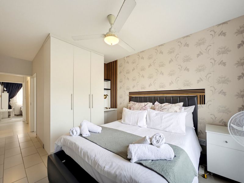 A well-maintained bedroom featuring built-in wardrobes, a ceiling fan, decorative wallpaper, and tiled flooring, offering a comfortable and functional living space.