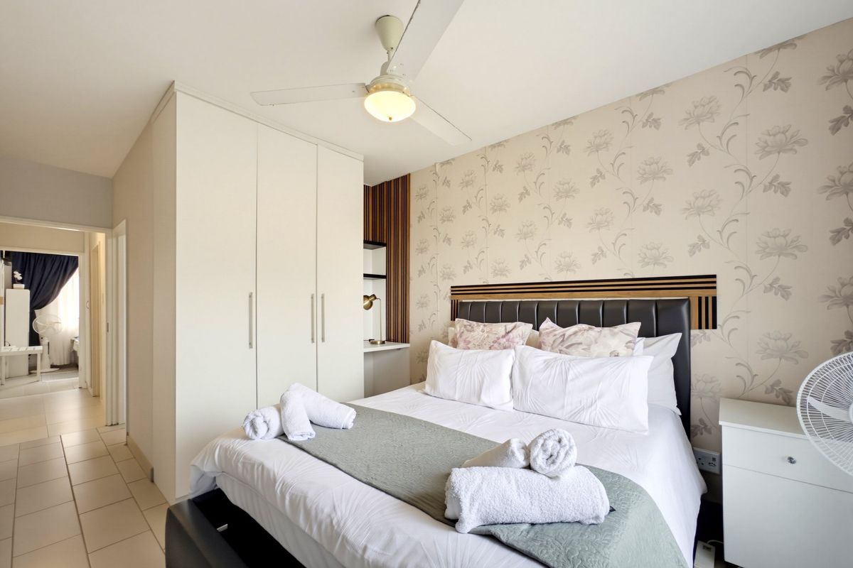 A well-maintained bedroom featuring built-in wardrobes, a ceiling fan, decorative wallpaper, and tiled flooring, offering a comfortable and functional living space.
