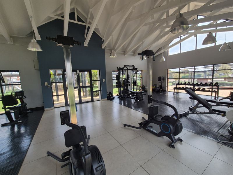 A spacious, well-lit gym facility featuring vaulted ceilings with exposed rafters, large windows, and a combination of tiled and rubber flooring.