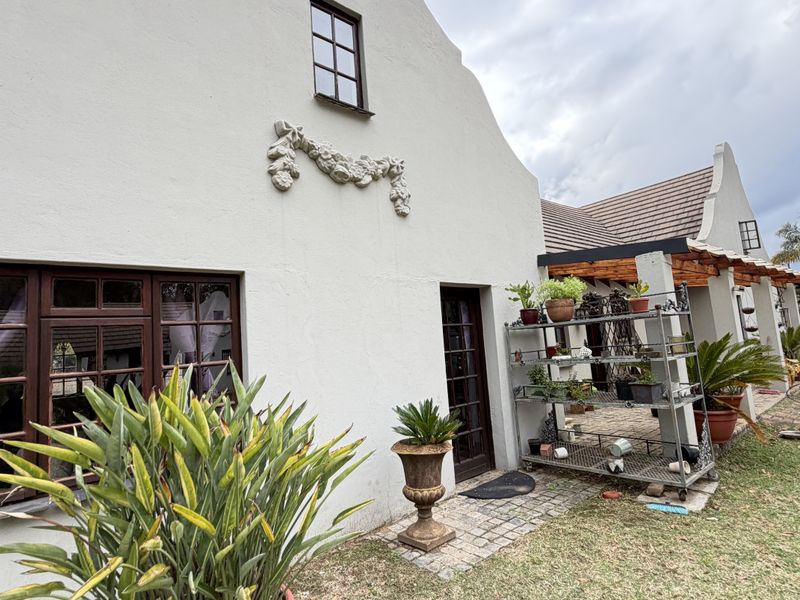 Rear elevation of a Cape Dutch style home featuring a gabled roof, decorative wall relief, timber frame windows, and a paved walkway under a pergola.