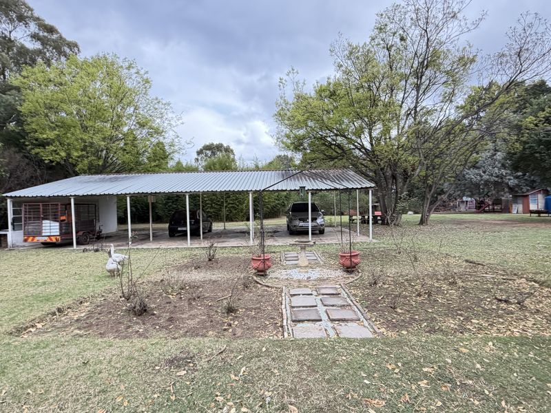 A spacious rear garden featuring a large corrugated metal carport structure, mature trees, and a stone pathway leading to a central garden feature.