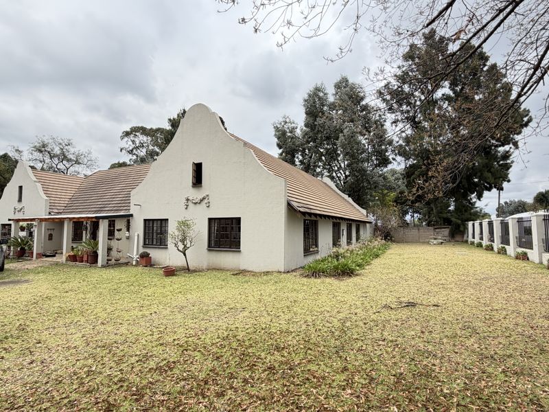 A Cape Dutch style house featuring a distinctive gable, tiled roof, and a large front garden with perimeter security fencing.
