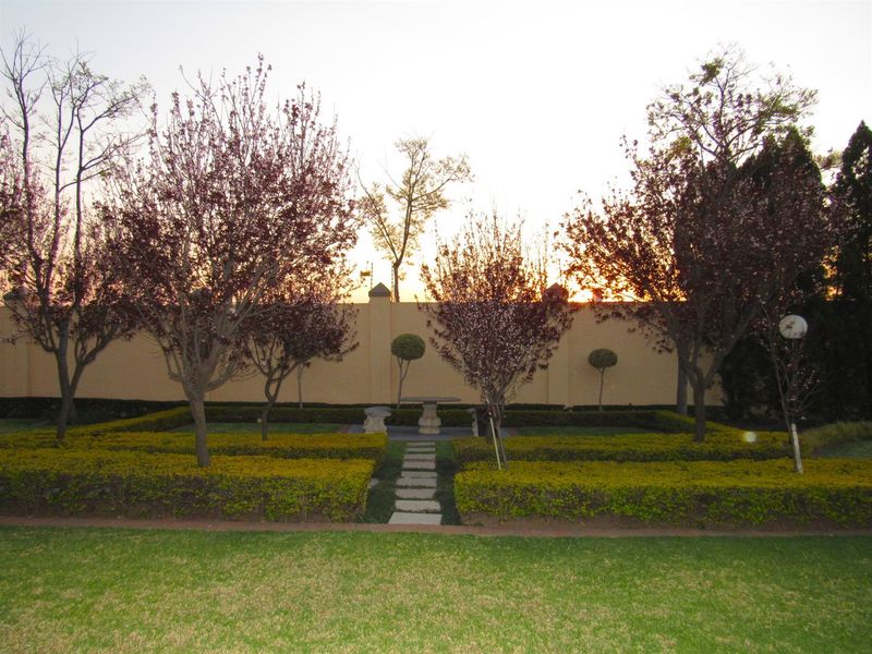 A well-maintained garden featuring manicured hedges, a stone pathway, and mature trees set against a perimeter wall at sunset.